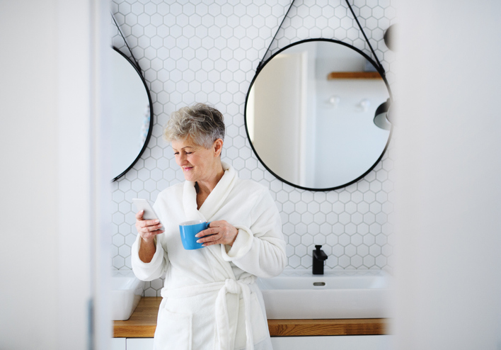 Active senior woman with bathrobe in bathroom, using smartphone.