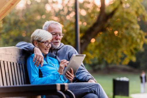 Smiling,Senior,Active,Couple,Sitting,On,The,Bench,Looking,At Two seniors looking at their iPad while sitting on a bench in a park