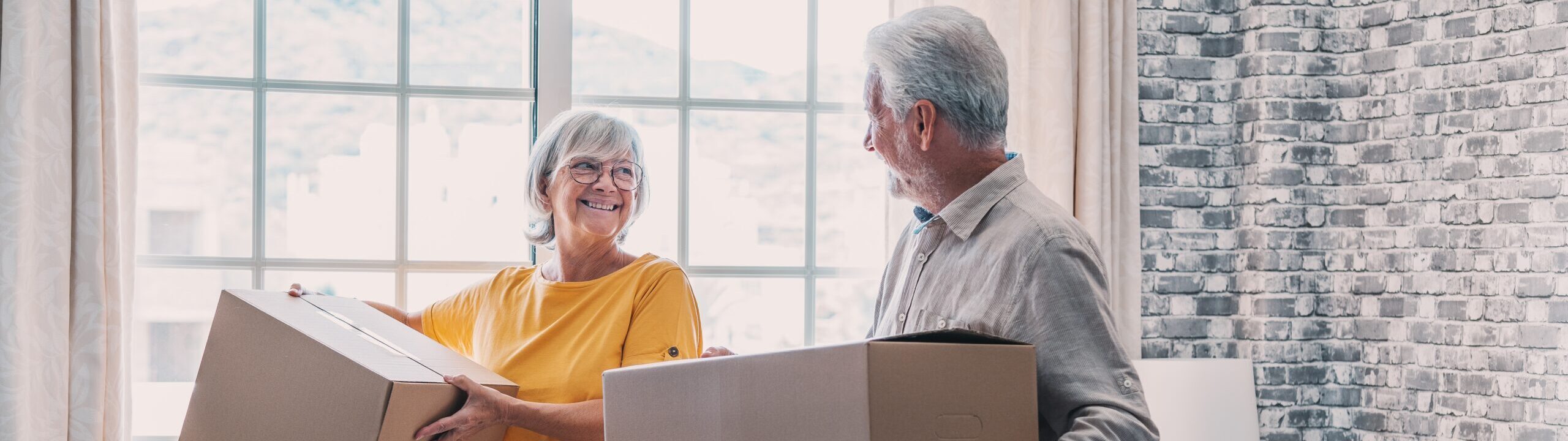 Mature couple moving into new apartment, carrying cardboard boxes into empty room with potted plants. Real estate property buying, relocation, new home concept. Rear view