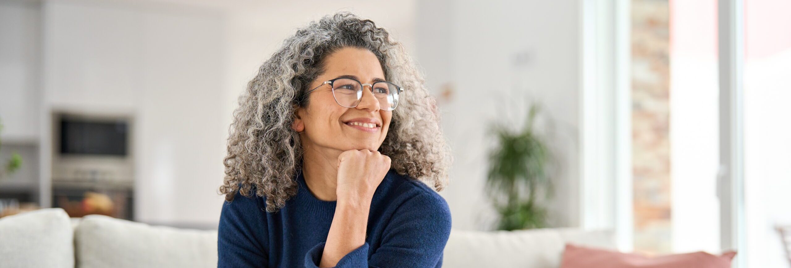 Happy relaxed 50 years old woman sitting on sofa at home looking away. Smiling middle aged lady posing in living room at home. Mature older female with curly gray hair relaxing on couch enjoying chill
